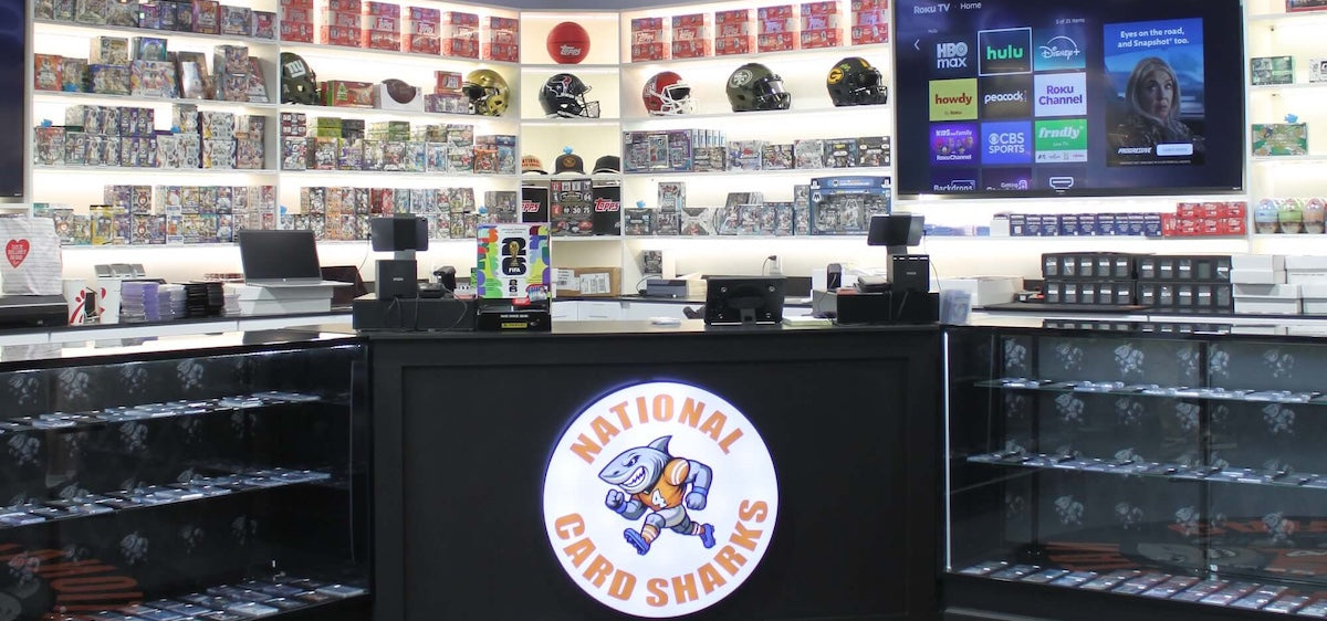 NCS Randolph store interior with a counter featuring a 'National Card Sharks' logo, shelves with products, and a TV screen.